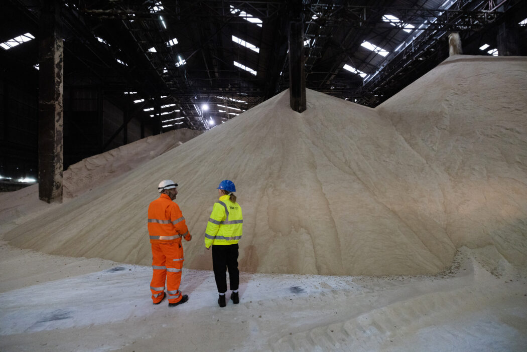 Young person talks to man in hi vis coat about sugar in front of pile of raw sugar