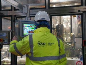 Man in hi vis jacket and hardhat at a factory looks at a computer screen