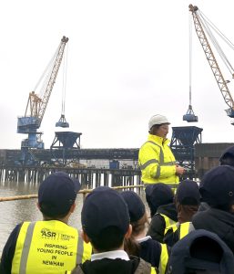 A group of students wearing navy caps and yellow high‑visibility vests listen to a person in a bright high‑visibility jacket and hard hat during an outdoor tour at an industrial waterfront site. Two large cranes and a long pier structure extend over the water in the background. The back of one student’s vest displays the Tate & Lyle Sugars logo and the words “Inspiring Young People in Newham.” Provide your feedback on BizChat