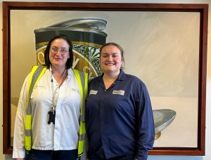 Two women, one in navy jumper and one in a white shirt and yellow hi-vis jacket stand smiling in front of a painting of a half-open can of Lyle's Golden Syrup