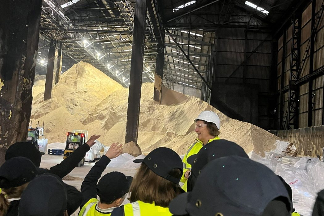 A group of students wearing navy caps and yellow high‑visibility vests listen to a woman in a bright high‑visibility jacket and hard hat during a tour in a warehouse. hard hat. In front of them are massive piles of raw sugar reaching nearly to the metal roof structure. Forklifts and machinery are visible near the base of the sugar stacks, and overhead lights illuminate the expansive interior.