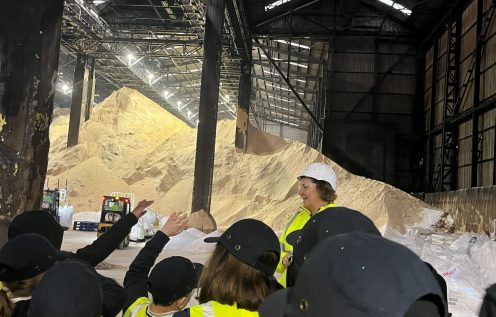 A group of students wearing navy caps and yellow high‑visibility vests listen to a woman in a bright high‑visibility jacket and hard hat during a tour in a warehouse. hard hat. In front of them are massive piles of raw sugar reaching nearly to the metal roof structure. Forklifts and machinery are visible near the base of the sugar stacks, and overhead lights illuminate the expansive interior.
