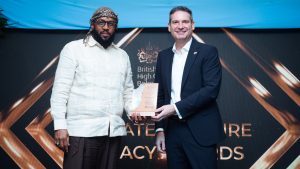 Two individuals stand side by side on a stage, holding an award plaque together. They are positioned in front of a large backdrop displaying the British High Commission logo and geometric gold patterns. One person is dressed in a light-colored long-sleeved shirt and dark trousers, while the other wears a dark suit with a white shirt. A blue banner hangs above them as they pose for a formal award presentation photograph.