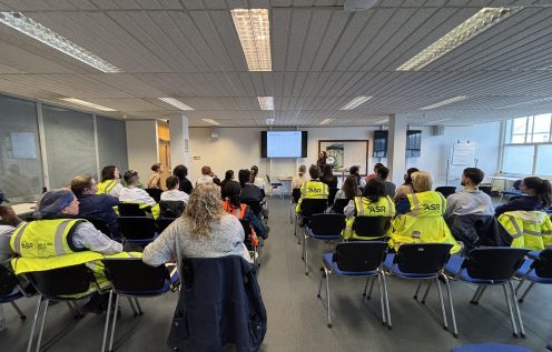 Office workers and workers in ASR hi-vis jackets sit in rows of chairs facing a screen that reads overcoming Imposter Syndrome and two female speakers