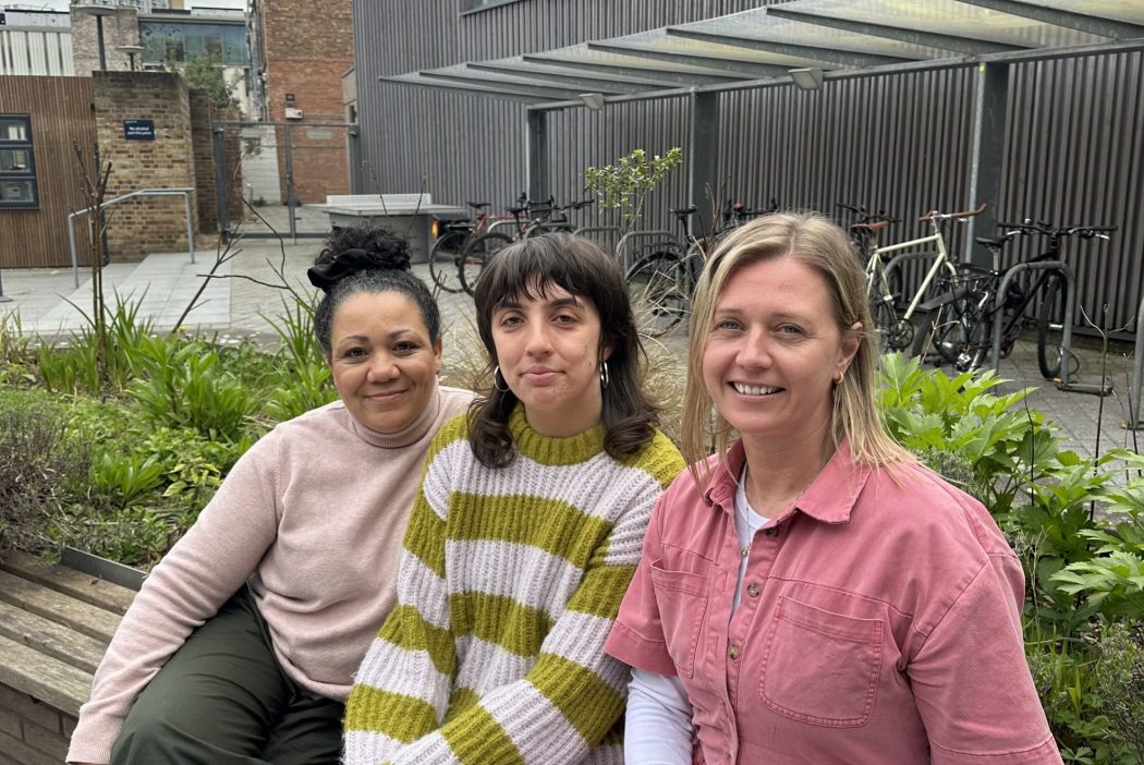 Three women sit on a bench outside a modern building