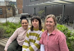 Three women sit on a bench outside a modern building