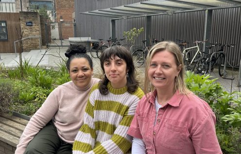Three women sit on a bench outside a modern building