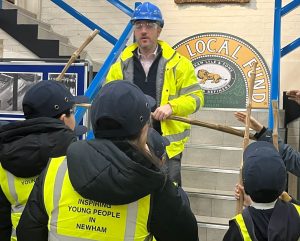 Children in hi-vis jackets that read "Inspiring Young People in Newham" face a man in hi-vis and a hard hat holding dry sugar cane. He stands in front of a staircase, at the top of which is a sign that reads "Local Fuind" with the Lyle's Golden Syrup logo beneath. Also in the background is a board with black and white pictures.