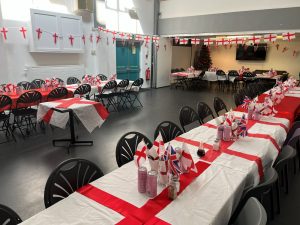 Long tables and chairs dressed with tablecloths with the St. George's flag on them. Laid out are cutlery, drinks, and little flags decorating the tables. Banners of the St. George's cross decorate the community centre's hall