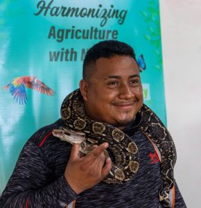 A Belizean man smiles, holding a snake around his snake. Behind him, a sign reads harmonising agriculture with nature