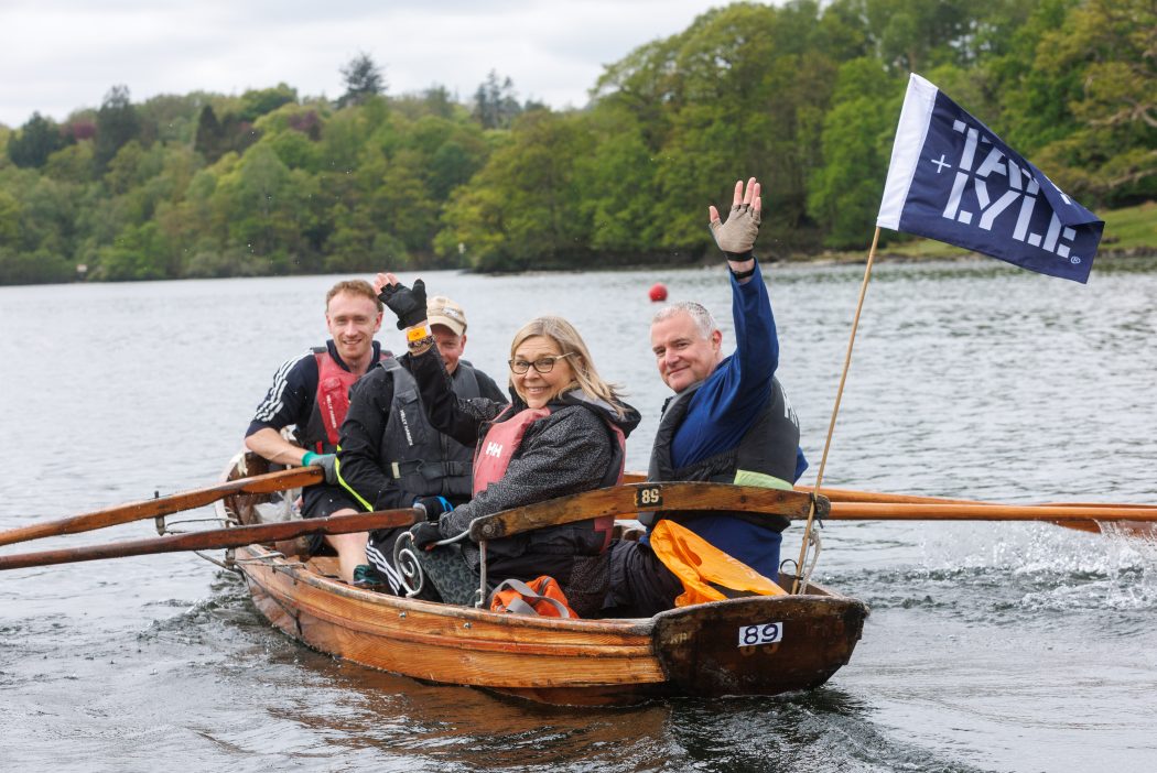 4 people wave from a row boat on a lake