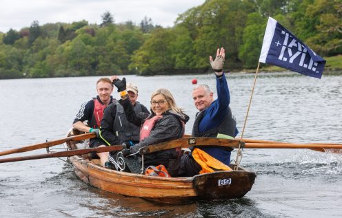 4 people wave from a row boat on a lake