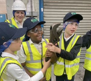 Three children together hold a large tool with an adult behind them