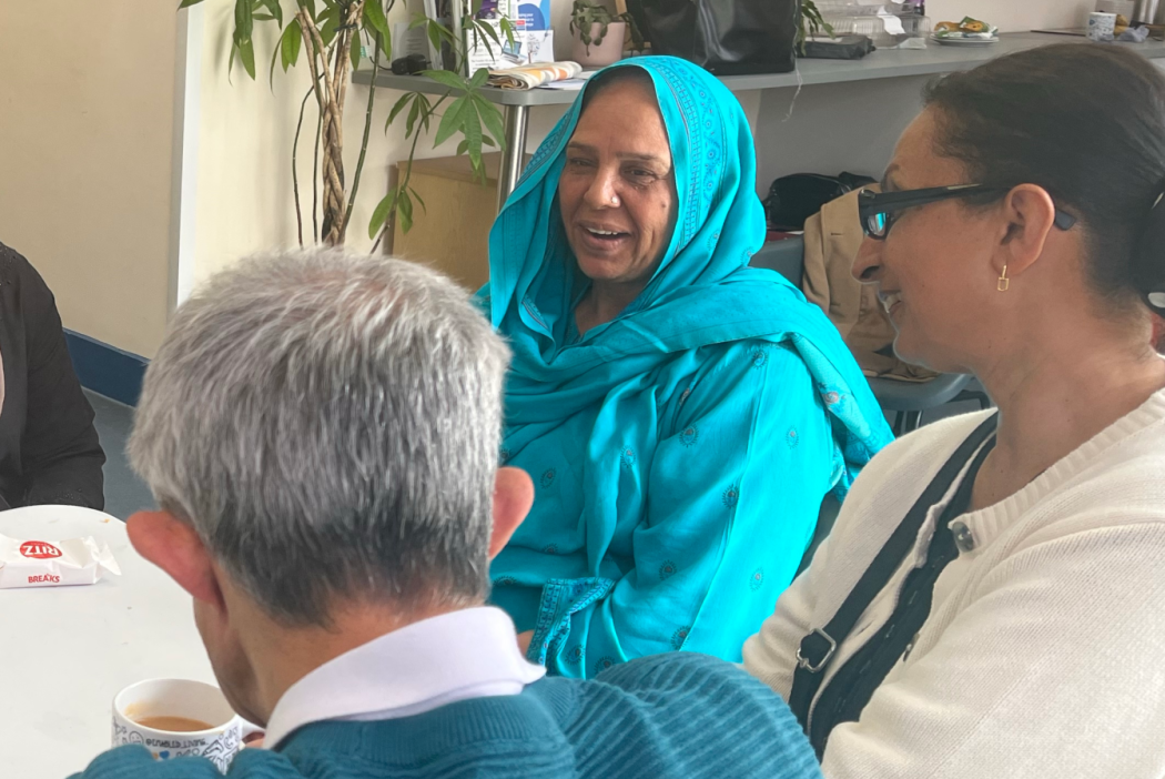 People gathered around a table at a community coffee morning, talking over tea and refreshments in a bright community centre meeting room.