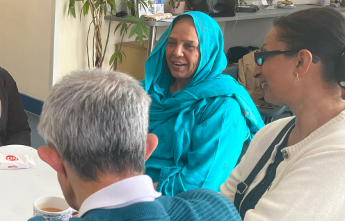 People gathered around a table at a community coffee morning, talking over tea and refreshments in a bright community centre meeting room.