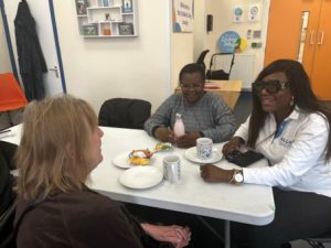 Three people chatting around a table during a community coffee morning. Mugs, drinks and snacks are on the table, and the event takes place in a bright meeting room.